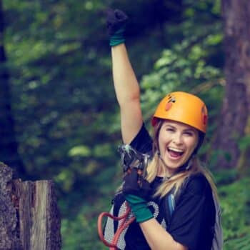 Fröhliche Frau mit Kletterausrüstung und Helm jubelt im Wald.