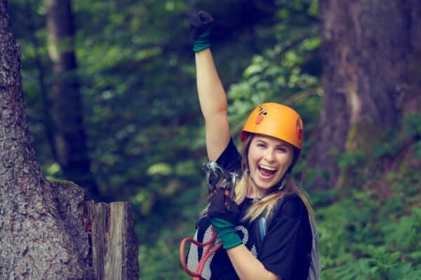 Fröhliche Frau mit Kletterausrüstung und Helm jubelt im Wald.