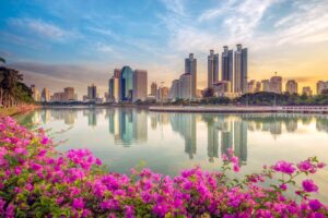 Skyline von Bangkok bei Sonnenaufgang mit Spiegelung im See und pinken Blumen im Vordergrund, Thailand