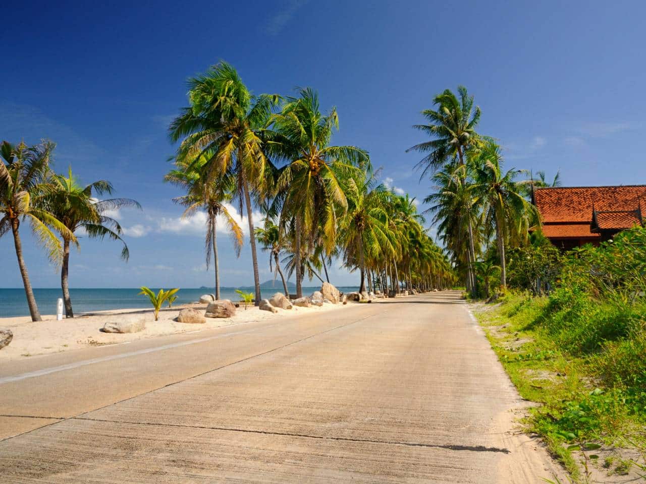 Leere Strandstraße in Ban Krut gesäumt von Palmen, links der Golf von Thailand, rechts ein traditionelles Haus mit rotem Dach