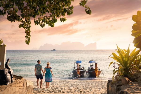 Paar hält Händchen am Strand bei Sonnenuntergang mit Longtail-Booten im Hintergrund